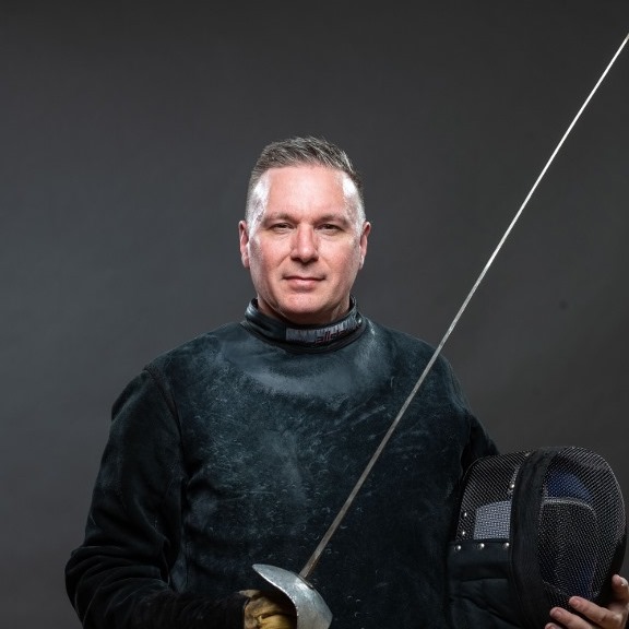 Tim Stang in fencing uniform holding a sword and mask
